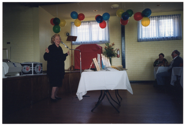 COL photo of a woman speaking into a microphone on a stand. She is standing behind a long rectangular table. On the table are stacks and displays of copies of books and two signs. In the background, on the stage behind her, there is a large unidentifiable contraption, a large cardboard box, and to the side of the stage, a tall wooden lectern, a red cover over a piano, and a floral bouquet in a tall vase. Next to the vase of flowers, there is a small fire extinguisher affixed to the wall. There are colourful balloons in red, yellow, blue, and green hanging from the ceiling. Two guests are sitting at a table, to the right of the frame. This photo has been taken from a side view of the table.