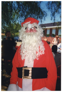 COL photo of a man, dressed in a Santa costume, standing beneath a large tree. Behind him, a woman is holding up a Photographic camera while others are seated or standing nearby. There is a red brick building in the background.