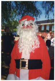 COL photo of a man, dressed in a Santa costume, standing beneath a large tree. Behind him, a woman is holding up a Photographic camera while others are seated or standing nearby. There is a red brick building in the background.