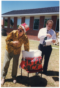 COL photo of a woman, wearing a Santa hat, reaching into a large red and white bag with the words, "Santa's Sack", in black lettering on the front. Beside her, a man is looking down at a piece of paper in his hand. They are standing on a grassy area outside a red brick building. In the background, there is a large dark green rubbish bin to the left of the frame, and a large hessian paper recycling bag on a metal frame to the right of the frame.