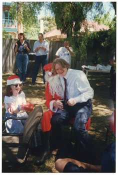 COL photo of a person dressed in a Santa costume sitting on a chair on a grassy area outdoors. A young man is sitting on his knee, laughing. A young girl, also laughing, is sitting beside them. She is wearing a red and white Santa hat and holding a gift-wrapped present. In the background, three people are standing near the fence, in front of a large green rubbish bin. Beside them, there is a long rectangular outdoor wooden table beneath a large tree. The table is covered in party plates and cups, amongst other miscellaneous items. In the background, there is a two-storey house and a one-storey house behind a wooden fence.