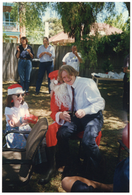 COL photo of a person dressed in a Santa costume sitting on a chair on a grassy area outdoors. A young man is sitting on his knee, laughing. A young girl, also laughing, is sitting beside them. She is wearing a red and white Santa hat and holding a gift-wrapped present. In the background, three people are standing near the fence, in front of a large green rubbish bin. Beside them, there is a long rectangular outdoor wooden table beneath a large tree. The table is covered in party plates and cups, amongst other miscellaneous items. In the background, there is a two-storey house and a one-storey house behind a wooden fence.