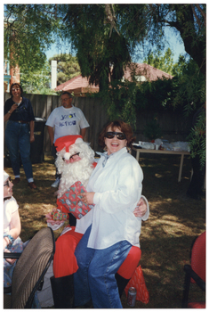 COL photo of a person dressed in a Santa costume sitting on a chair on a grassy area outdoors. A woman is sitting on his knee, laughing. A young girl (partially obscured) is sitting beside them. In the background, two people are standing near the fence, in front of a large green rubbish bin. Beside them, there is a long rectangular outdoor wooden table beneath a large tree. The table is covered in party plates and cups, amongst other miscellaneous items. In the background, there is a two-storey house and a one-storey house behind a wooden fence.