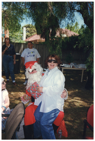 COL photo of a person dressed in a Santa costume sitting on a chair on a grassy area outdoors. A woman is sitting on his knee, laughing. A young girl (partially obscured) is sitting beside them. In the background, two people are standing near the fence, in front of a large green rubbish bin. Beside them, there is a long rectangular outdoor wooden table beneath a large tree. The table is covered in party plates and cups, amongst other miscellaneous items. In the background, there is a two-storey house and a one-storey house behind a wooden fence.