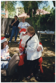 COL photo of a person dressed in a Santa costume sitting on a chair on a grassy area outdoors. A woman is sitting on his knee, facing him. A young girl is sitting beside them. She is wearing a red and white Santa hat. In the background, two people are standing near the fence, in front of a large green rubbish bin. Beside them, there is a long rectangular outdoor wooden table beneath a large tree. The table is covered in party plates and cups, amongst other miscellaneous items. In the background, there is a two-storey house and a one-storey house behind a wooden fence.