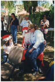 COL photo of a person dressed in a Santa costume sitting on a chair on a grassy area outdoors. A woman is sitting on his knee, smiling at a girl sitting beside them. The girl is wearing a red and white Santa hat. In the background, four people are standing near the fence, in front of a large green rubbish bin. One man is eating a filled bread roll, which he is holding in one hand. There are two women standing beside him, each holding a drink in their hand. On his other side, there is another woman eating from a plate, while standing. Behind her, there is a long rectangular outdoor wooden table beneath a large tree. The table is covered in party plates and cups, amongst other miscellaneous items. In the background, there is a two-storey house and a one-storey house behind a wooden fence.
