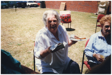 COL medium close-up photo of two women sitting on chairs outdoors. One woman is holding a plate of barbequed meat with a drink cup in one hand, and a serviette in the other. She is facing the camera, laughing. The other woman (partially obscured) is holding a cup of orange juice in her hand. Behind them, there is a big red and white bag on a chair, with "Santa's Sack" in black lettering on the front. In the background, there is a red brick building and some cars parked in an adjacent car park. Against the exterior wall of the building, there is a large dark green rubbish bin, a small wooden pallet leaning against the brick wall, and a large hessian paper recycling bag on a metal frame.
