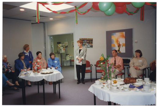 COL photo of a woman standing in a large room, while several others are seated around the room. In the foreground, there are two hexagonal tables covered in white tablecloths. On top of the tables, there are plates of food, cups and saucers, a stack of small plates, a floral arrangement on one table and a decorated pine tree branch on the other. There are green and red balloons and streamers hanging from the ceiling. There are posters on the wall and on a display board. Through an open double doorway, a tall stand with brochures is evident, with a smaller stand filled with brochures on a small table beneath it. There is a burgundy red chair next to the small table, and some posters on the wall.