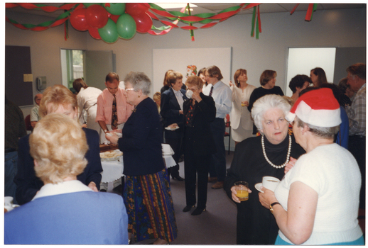COL photo of a large group of people standing around a room, eating and drinking. Amongst the crowd, there is a table with various food platters on top. There are green and red balloons and streamers hanging from the ceiling.