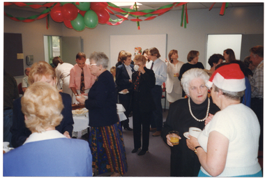 COL photo of a large group of people standing around a room, eating and drinking. Amongst the crowd, there is a table with various food platters on top. There are green and red balloons and streamers hanging from the ceiling.