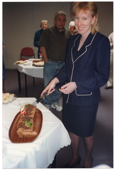 COL photo of a woman holding a large knife poised over a chocolate yule log (a cake in the shape of a log). The yule log has been decorated with some plastic holly leaves and berries, and a Santa figurine. There is also a plate of party pies on the table. In the background, there is another table with various food platters on top. A few people are standing in the background, looking on.