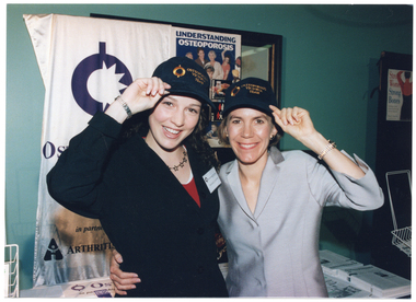 COL medium close-up photo of two women standing with their arms around each other. They are both wearing navy blue caps embroidered at the front in gold with, "Osteoporosis Victoria Team", each touching the peak of their own cap with one hand. Behind them, there is a table with various printed information sheets on top. On the wall, there is a banner with the text, "Osteoporosis Victoria in partnership with Arthritis Victoria" (partially obscured), and a framed noticeboard with a poster on it. There is a "Strong Women, Strong Bones" poster on an adjacent wall.