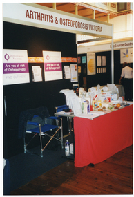 COL photo of an information stand at an expo. On a rectangular table covered with a red tablecloth, there is a range of printed factsheets arranged on display stands, and fresh and packaged food items with labels attached. At the top of the information booth, there is a sign that says, "ARTHRITIS & OSTEOPOROSIS VICTORIA". Behind the table, there are posters, brochures, and information sheets on a dividing wall separating the stalls. The text on the poster says, "OSTEOPOROSIS VICTORIA Are you at risk of Osteoporosis? FIND OUT HERE". There are also two blue deck chairs behind the table. Another booth is evident in the background.