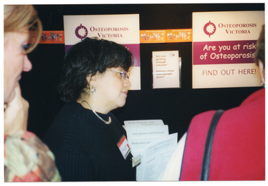 COL close-up photo of three women (two partially obscured) at an information stand. Headings on some printed factsheets on a display stand include "Bone Density and Osteoporosis", "Osteoporos and Excercise", and "Medications and..." (partially obscured). at an information stall. Two posters on the wall contain the text, "OSTEOPOROSIS VICTORIA Are you at risk of Osteoporosis? FIND OUT HERE". Between the posters, a small sign with the "OV" logo (for Osteoporosis Victoria) says, "Are you getting enough calcium? Surveys show most people don't!"