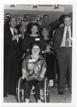 B&W photo of a group of people posing for a photo. Front and centre is a woman dressed in a sequinned top and satin trousers. She is a wheelchair user. Behind her, there is a small group of people, standing. Two of them are holding champagne flutes in one hand.