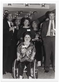 B&W photo of a group of people posing for a photo. Front and centre is a woman dressed in a sequinned top and satin trousers. She is a wheelchair user. Behind her, there is a small group of people, standing. Two of them are holding champagne flutes in one hand.