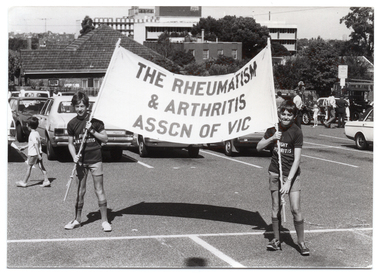 B&W photo of two boys each holding up a wooden pole onto which a large banner is attached. They are in a carpark, with some houses, trees, and multistorey buildings evident in the background. Both boys are wearing a t-shirt with the text, "I FIGHT ARTHRITIS", and shorts. The text on the banner says, "THE RHEUMATISM & ARTHRITIS ASSCN OF VIC". There are several cars parked in the car park, including two vintage cars to the right of the frame. A few people are looking at the vintage cars.