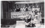 B&W photo of a group of people standing on the corner veranda of a building, and a few others sitting on a garden bench or standing in front of the veranda. There is a set of large windows on each of the two walls surrounding the veranda. A large banner hanging from the metal ballustrade across the front of the veranda contains the text, "Arthritis Foundation of Victoria Ph: 03 862-2555 EXERCISE HELPS BEAT ARTHRITIS Funded by Vic Health Promotion Foundation".