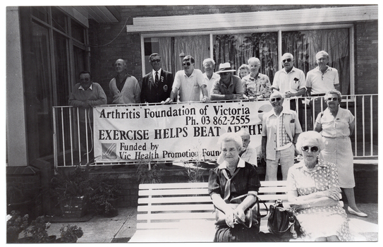 B&W photo of a group of people standing on the corner veranda of a building, and a few others sitting on a garden bench or standing in front of the veranda. There is a set of large windows on each of the two walls surrounding the veranda. A large banner hanging from the metal ballustrade across the front of the veranda contains the text, "Arthritis Foundation of Victoria Ph: 03 862-2555 EXERCISE HELPS BEAT ARTHRITIS Funded by Vic Health Promotion Foundation".