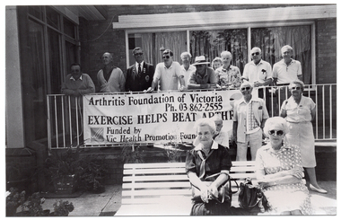 B&W photo of a group of people standing on the corner veranda of a building, and a few others sitting on a garden bench or standing in front of the veranda. There is a set of large windows on each of the two walls surrounding the veranda. A large banner hanging from the metal ballustrade across the front of the veranda contains the text, "Arthritis Foundation of Victoria Ph: 03 862-2555 EXERCISE HELPS BEAT ARTHRITIS Funded by Vic Health Promotion Foundation".