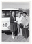 B&W photo of three women standing in front of a van. Two of the women are each holding a walking stick, one of whom is also holding a handbag. Another vehicle (partially obscured) and some small buildings are evident in the background.
