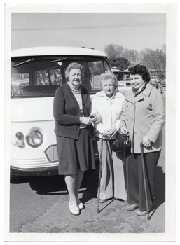 B&W photo of three women standing in front of a van. Two of the women are each holding a walking stick, one of whom is also holding a handbag. Another vehicle (partially obscured) and some small buildings are evident in the background.