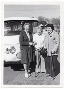 B&W photo of three women standing in front of a van. Two of the women are each holding a walking stick, one of whom is also holding a handbag. Another vehicle (partially obscured) and some small buildings are evident in the background.
