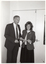 B&W photo of a man and a woman both holding a small piece of paper. They are both looking at the camera and smiling. There is a closed double door behind them.