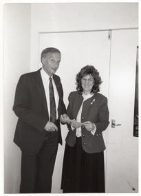 B&W photo of a man and a woman both holding a small piece of paper. They are both looking at the camera and smiling. There is a closed double door behind them.