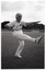 B&W photo of a woman, wearing a "Joint Action" t-shirt, doing some stretching exercises. She is holding one arm outstretched in front of her, while at the same time holding her opposite leg outstretched in front of her. She has her weight on one leg, and has her opposite hand (in a fist) on her hip. There is a large grassy sports ground in the background, with a metal goal frame in the distance.