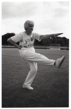 B&W photo of a woman, wearing a "Joint Action" t-shirt, doing some stretching exercises. She is holding one arm outstretched in front of her, while at the same time holding her opposite leg outstretched in front of her. She has her weight on one leg, and has her opposite hand (in a fist) on her hip. There is a large grassy sports ground in the background, with a metal goal frame in the distance.
