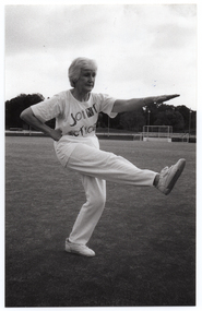 B&W photo of a woman, wearing a "Joint Action" t-shirt, doing some stretching exercises. She is holding one arm outstretched in front of her, while at the same time holding her opposite leg outstretched in front of her. She has her weight on one leg, and has her opposite hand (in a fist) on her hip. There is a large grassy sports ground in the background, with a metal goal frame in the distance.