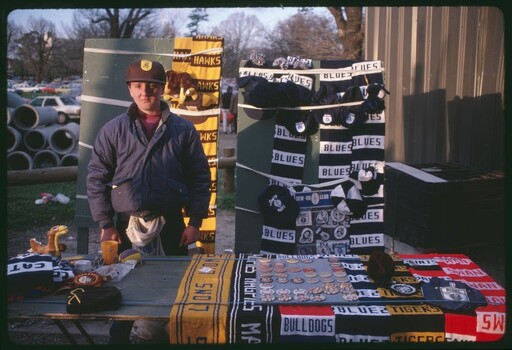 Photograph of a man standing with table of football fans’ merchandise