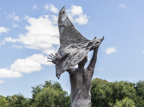 A statue of a naked man waving a piece of cloth throwing his hands up in the air. The whole statue including the cloth is cast in bronze.