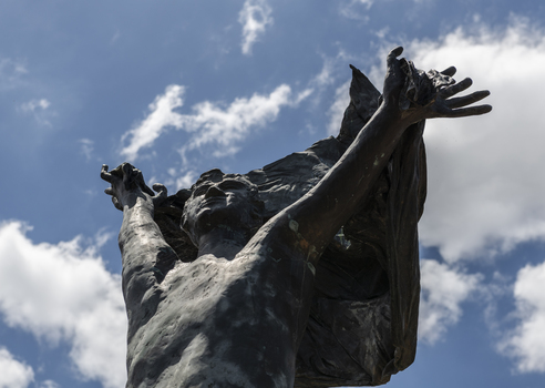 A statue of a naked man waving a piece of cloth throwing his hands up in the air. The whole statue including the cloth is cast in bronze.