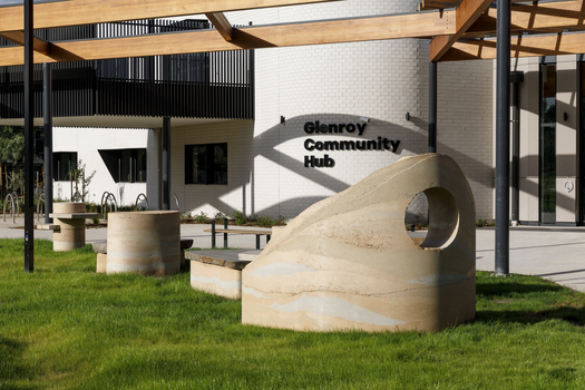 A large abstracted sculpture made out of stone that resembles a play structure structure or outdoor seating. it is in a grassy part out the front of a community hub. 