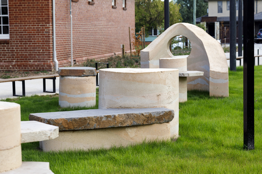A large abstracted sculpture made out of stone that resembles a play structure structure or outdoor seating. it is in a grassy part out the front of a community hub. 
