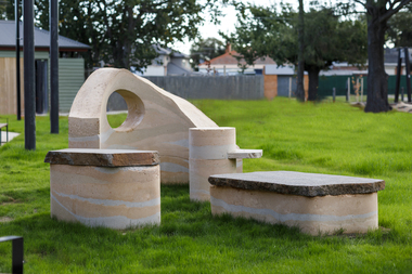 A large abstracted sculpture made out of stone that resembles a play structure structure or outdoor seating. it is in a grassy part out the front of a community hub. 