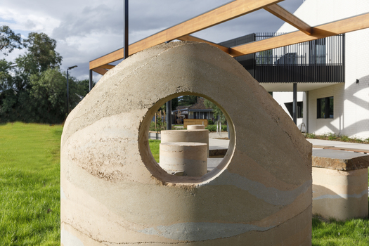 A large abstracted sculpture made out of stone that resembles a play structure structure or outdoor seating. it is in a grassy part out the front of a community hub. 