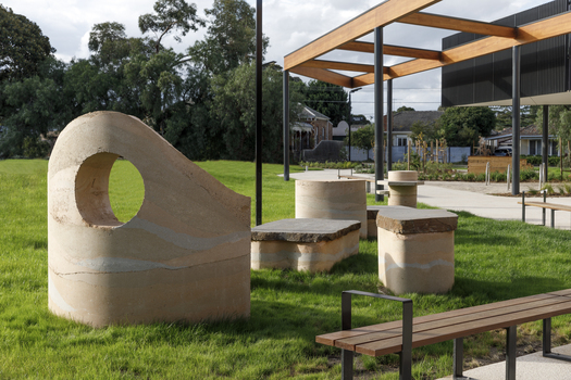 A large abstracted sculpture made out of stone that resembles a play structure structure or outdoor seating. it is in a grassy part out the front of a community hub. 