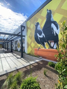 An outdoor mural featuring 2 larger larger than life magpies. they are sitting in front of a geometric pattern.