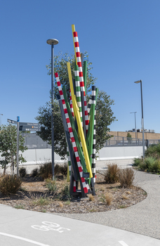 A large sculpture consisting of a series of striped and unstriped metal poles sticking out of the ground in a single cluster in a haphazard manner. there are approximately 15 of them and the sculpture is located on a street corner.