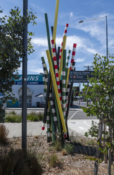 A large sculpture consisting of a series of striped and unstriped metal poles sticking out of the ground in a single cluster in a haphazard manner. there are approximately 15 of them and the sculpture is located on a street corner.