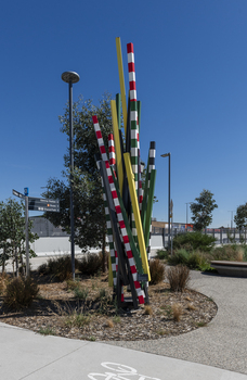 A large sculpture consisting of a series of striped and unstriped metal poles sticking out of the ground in a single cluster in a haphazard manner. there are approximately 15 of them and the sculpture is located on a street corner.