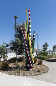 A large sculpture consisting of a series of striped and unstriped metal poles sticking out of the ground in a single cluster in a haphazard manner. there are approximately 15 of them and the sculpture is located on a street corner.