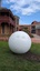 A large metal sphere sculpture with abstract butterfly motifs cut into it. It is located in a garden in front of a building.