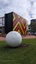 A large metal sphere sculpture with abstract butterfly motifs cut into it. It is located in a garden in front of a building featuring a geometric abstract pattern.