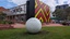 A large metal sphere sculpture with abstract butterfly motifs cut into it. It is located in a garden in front of a building featuring a geometric abstract pattern.
