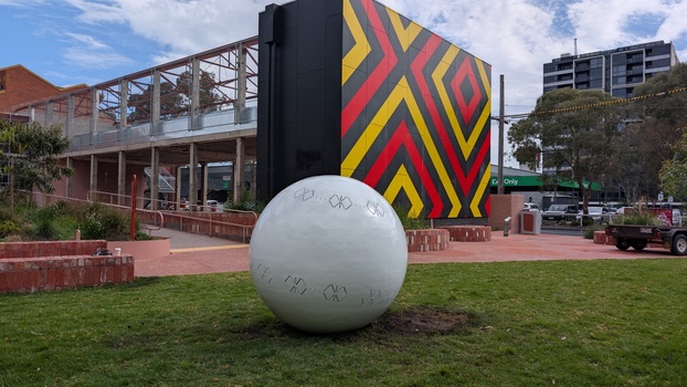 A large metal sphere sculpture with abstract butterfly motifs cut into it. It is located in a garden in front of a building featuring a geometric abstract pattern.