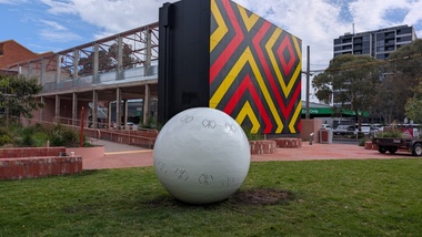 A large metal sphere sculpture with abstract butterfly motifs cut into it. It is located in a garden in front of a building featuring a geometric abstract pattern.